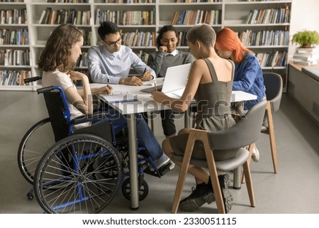 Diverse group of students inclusively college mate with disability sitting at table in library together, writing notes, working on creative essay, doing school homework together. Full length Royalty-Free Stock Photo #2330051115