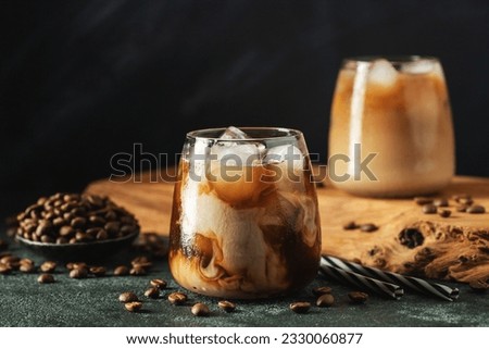 Ice coffee in a tall glass with cream poured over, ice cubes and beans on a dark concrete table. Royalty-Free Stock Photo #2330060877