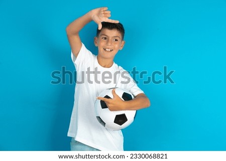 Little hispanic boy wearing white T-shirt holding a football ball making finger frame with hands. Creativity and photography concept.