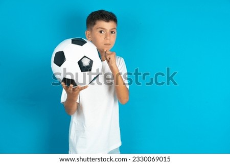 Little hispanic boy wearing white T-shirt holding a football ball Ready to fight with fist defense gesture, angry and upset face, afraid of problem.
