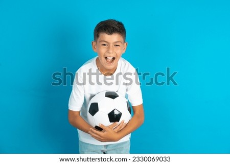 Little hispanic boy wearing white T-shirt holding a football ball  smiling and laughing hard out loud because funny crazy joke with hands on body.