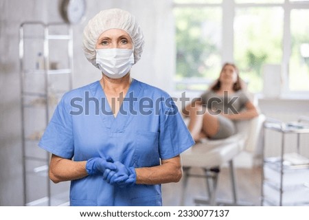 Portrait of elderly female doctor wearing uniform, medical mask and gloves standing in treatment room at beauty clinic
