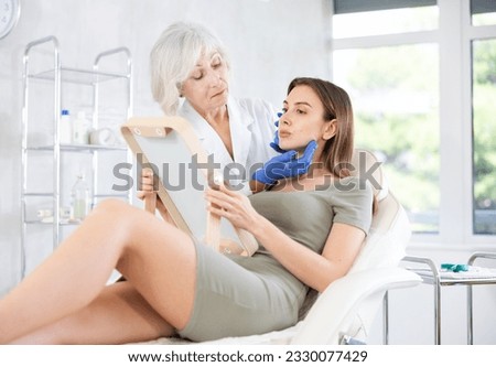Mature female cosmetologist consulting her young long haired female patient sitting in front of mirror at treatment room .