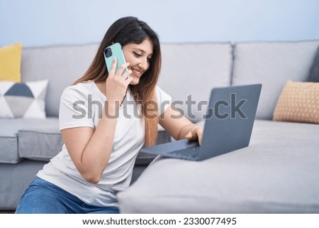 Young hispanic woman using laptop talking on the smartphone at home