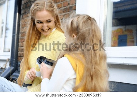 Mother and daughter sit outside and drink hot chocolate. The mother looks at her daughter with love and smiles. Family spending the weekend together. Horizontal photo