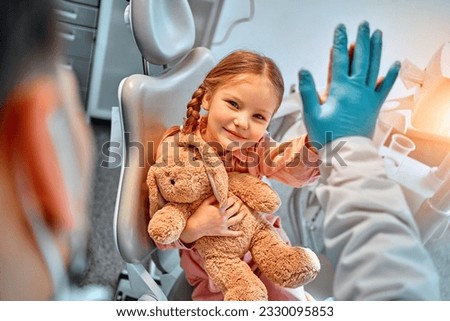 Portrait of a cute little girl with a toy rabbit on her lap sitting in a dentist's chair, giving a high five to the doctor and laughing. Dental care, trust and patient care. Children's dentistry. Royalty-Free Stock Photo #2330095853