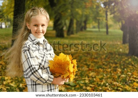 Little blonde girl posing in a white plaid shirt in an autumn park with a bouquet of yellow leaves. Horizontal photo