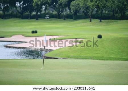 A golf ball that almost hits the hole with the flag. On the green with many signs of repair In the background are sand bunkers, ponds and lush lawns. There is a blurred golf cart in the distance. 