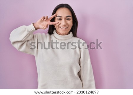 Young south asian woman standing over pink background doing peace symbol with fingers over face, smiling cheerful showing victory 