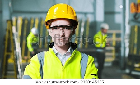 Portrait of beautiful Caucasian woman in helmet and uniform standing at plant manufactoring and taking on goggles. Indoors. Female worker at factory in hardhat and protectional glasses. Protection. Royalty-Free Stock Photo #2330145355