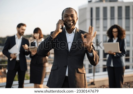 Beaming african american gentleman having conversation on smartphone during meeting with partners on terrace. Multiracial colleagues in suits using remote gadgets, showing sign ok