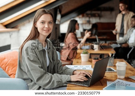 Young smiling female analyst or economist looking at camera while sitting by workplace in front of laptop and working with online financial data Royalty-Free Stock Photo #2330150779