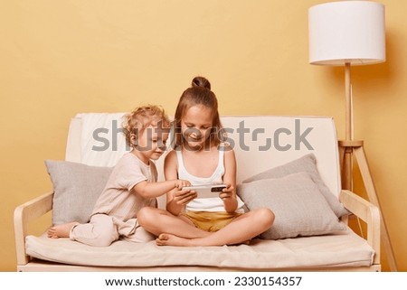 Positive playful little girls sisters sitting on couch using smartphone together posing against beige wall watching online video or cartoons expressing happiness.