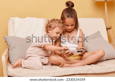 Concentrated children little girls sisters sitting on couch using smartphone together posing against beige wall playing on mobile phone looking at device display.