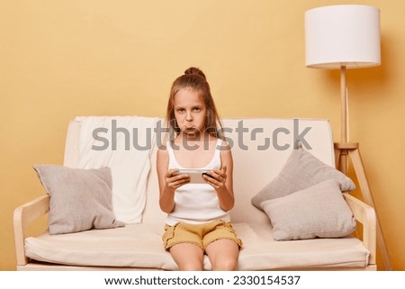 Sad upset little girl wearing casual attire sitting on sofa against beige wall using cell phone loosing game level looking at camera with pout lips expressing sadness.