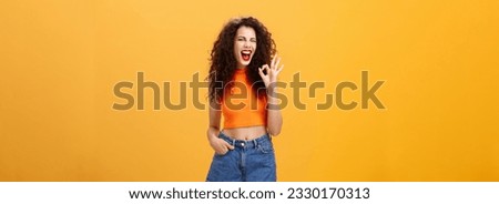 Excellent party like it. Joyful enthusiastic young curly-haired female with red lipstick in stylish cropped top winking excited smiling and showing okay or perfect sign posing over orange background.