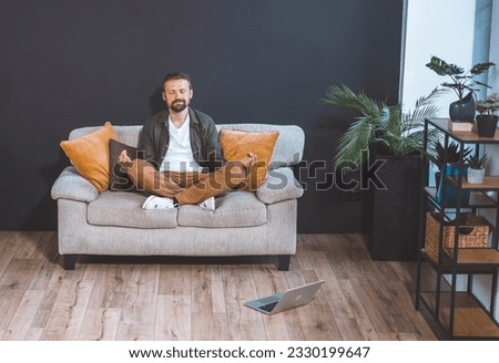 Man sitting home on couch, adopting lotus asana pose, and embracing state of relaxation. With serene expression on face, he immerses himself in practice of meditation or mindfulness. High quality Royalty-Free Stock Photo #2330199647