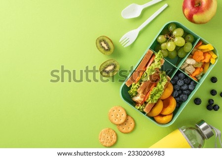 Lunch in the school cafeteria concept. Above view photo of lunch box with sandwiches, fruits and vegetables and water bottle on light green isolated background with copyspace for text or advert Royalty-Free Stock Photo #2330206983