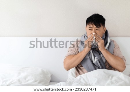 Sick unhealthy middle aged asian man wearing pajamas and scarf around his neck sitting in bed, sneezing, suffering from cold, flu, headache, runny nose, coronavirus, looking at copy space Royalty-Free Stock Photo #2330237471