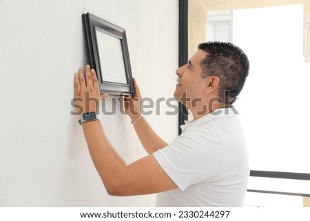 Dark-haired 40-year-old Latino man decorates his new apartment that he bought with a real estate agency, hangs a frame on the wall