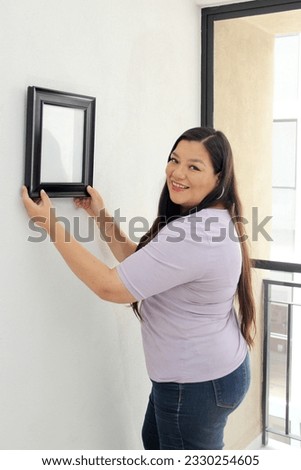 40-year-old Latina woman decorates her new apartment that she bought with a real estate agency, hangs a frame on the wall