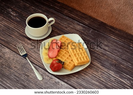 A cup of black coffee on a saucer, a fork and a plate with fresh viennese waffles with strawberries and bananas on a wooden table. Top view, flat lay.
