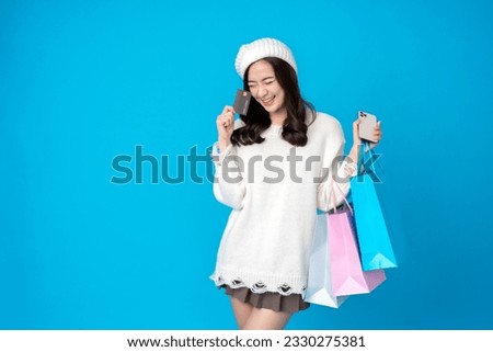 Asian female retailer with long hair holding credit card and smartphone for shopping and paper bag, wearing hat and white dress, smiling happily. Taking pictures in the blue background at the studio