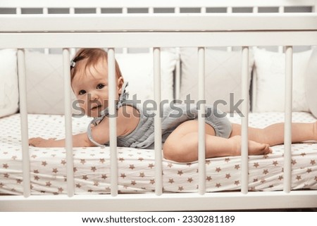 Happy little 9 month old girl lying on her stomach in her crib. Children's furniture, children's textiles. High quality photo Royalty-Free Stock Photo #2330281189