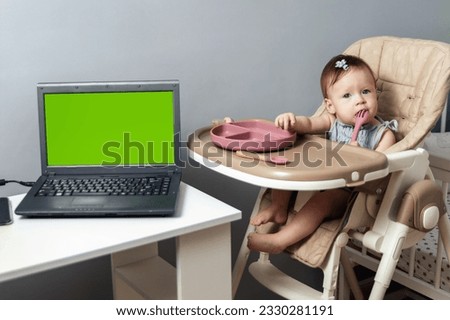Little gray-eyed 9-month-old girl sitting next to a laptop with a green screen. Hungry child waiting for lunch. The concept of healthy baby food. High quality photo