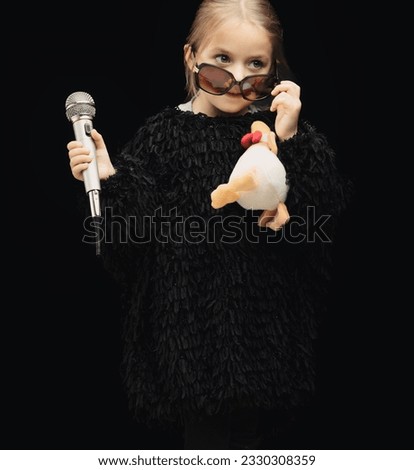 A girl dressed as a pop-rock star checks her audience, lowering her sunglasses. She holds a stuffed duck toy and a microphone. She might prefer being a social media influencer someday