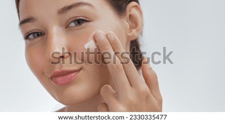 Face skin care portrait photo of a beautiful woman applying moisturizer cream on her face. Photo of smiling woman with perfect makeup on blue background. Beauty concept. High-quality photo