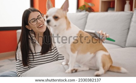 Young hispanic woman with chihuahua dog sitting on the floor brushing dog hair at home