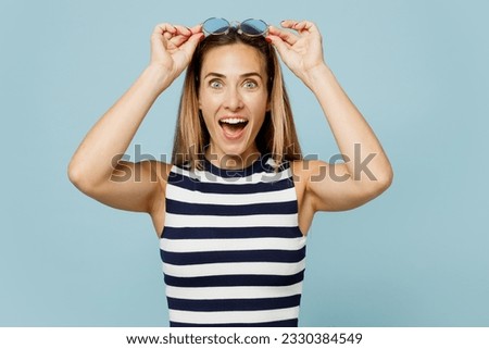 Young surprised fun blonde beautiful woman wear striped tank shirt casual clothes take off sunglasses look camera isolated on plain pastel light blue cyan background studio portrait. Lifestyle concept