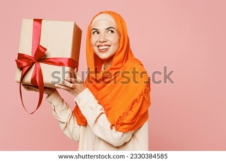 Sideways young arabian asian muslim woman wears orange abaya hijab hold present box with gift ribbon bowisolated on plain pink background studio portrait. Uae middle eastern islam religious concept