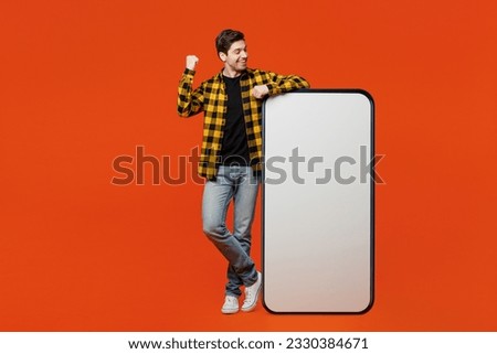 Full body excited young man wear yellow checkered shirt black t-shirt big huge blank screen mobile cell phone smartphone with workspace area do winner gesture isolated on plain red orange background