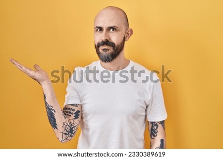 Young hispanic man with tattoos standing over yellow background smiling cheerful presenting and pointing with palm of hand looking at the camera. 