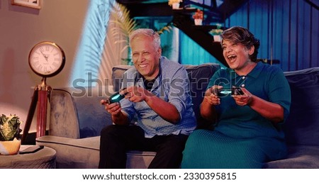 Happy Indian two excited smiling old pensioner couple playing video game hold gamepad in hand sitting on sofa having funny competition moment spend weekend time enjoy together late night indoor home Royalty-Free Stock Photo #2330395815