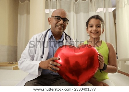 Indian happy little preschool girl and male doctor showing heart balloon Smiling small patient celebrating successful treatment finish with general practitioner at checkup. Royalty-Free Stock Photo #2330401599