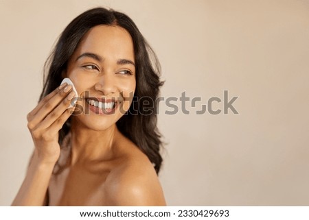 Beautiful young multiethnic woman with bare shoulder removing make up with cotton pad on cheek isolated against beige background. Smiling hispanic girl applying dry powder using cosmetic cushion. Royalty-Free Stock Photo #2330429693