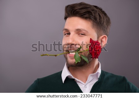 Half-length portrait of young handsome man wearing green sweater and white shirt keeping wonderful red rose in his mouth. Isolated on grey background Royalty-Free Stock Photo #233047771
