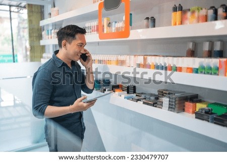 male vape store keeper calling on phone while checking the liquid stock using the digital tablet