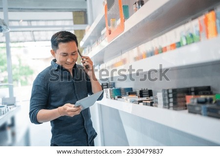 male shopkeeper standing beside the store shelf while calling on phone and looking at the digital tablet