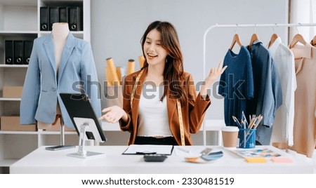 Asian Fashion designer woman talking smart phone and using laptop with digital tablet computer in modern studio the clothes hanging on the racks morning light
