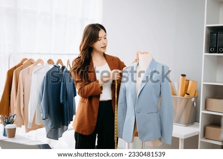 Asian Fashion designer woman talking smart phone and using laptop with digital tablet computer in modern studio the clothes hanging on the racks morning light