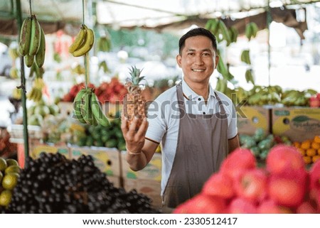 male seller in apron holding the pineapple at the fruit shop Royalty-Free Stock Photo #2330512417