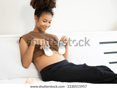 Happy multiracial pregnant woman holding baby shoe waiting for her unborn child. Young beautiful expectant mother relaxing on bed at home. Pregnancy and motherhood anticipation lifestyle.