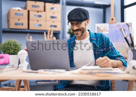Hispanic man with beard doing online video call at art studio doing ok sign with fingers, smiling friendly gesturing excellent symbol 