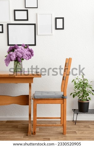 Vase with beautiful lilac flowers on table, chair and blank pictures in interior of light living room
