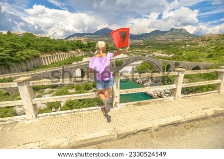 A woman visiting Mesi Bridge with Albania flag. Mesi Bridge is an Ottoman-era bridge located in the village of Mes, Albania. Largest in Albania from the Ottoman period, constructed in the 18th century Royalty-Free Stock Photo #2330524549
