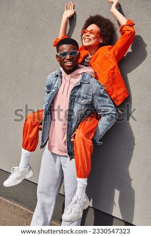 Positive and trendy young african american man in denim jacket holding best friend in sunglasses and bright outfit and standing near building on urban street, trendy friends in urban settings Royalty-Free Stock Photo #2330547323